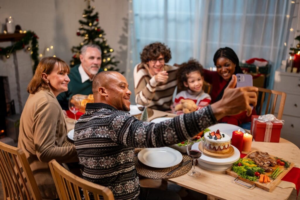 Multi-ethnic family using mobile phone video call online to relative. Attractive diverse group of people having dinner eating food to celebrate holiday Thanksgiving, X-mas eve on dining table at home.