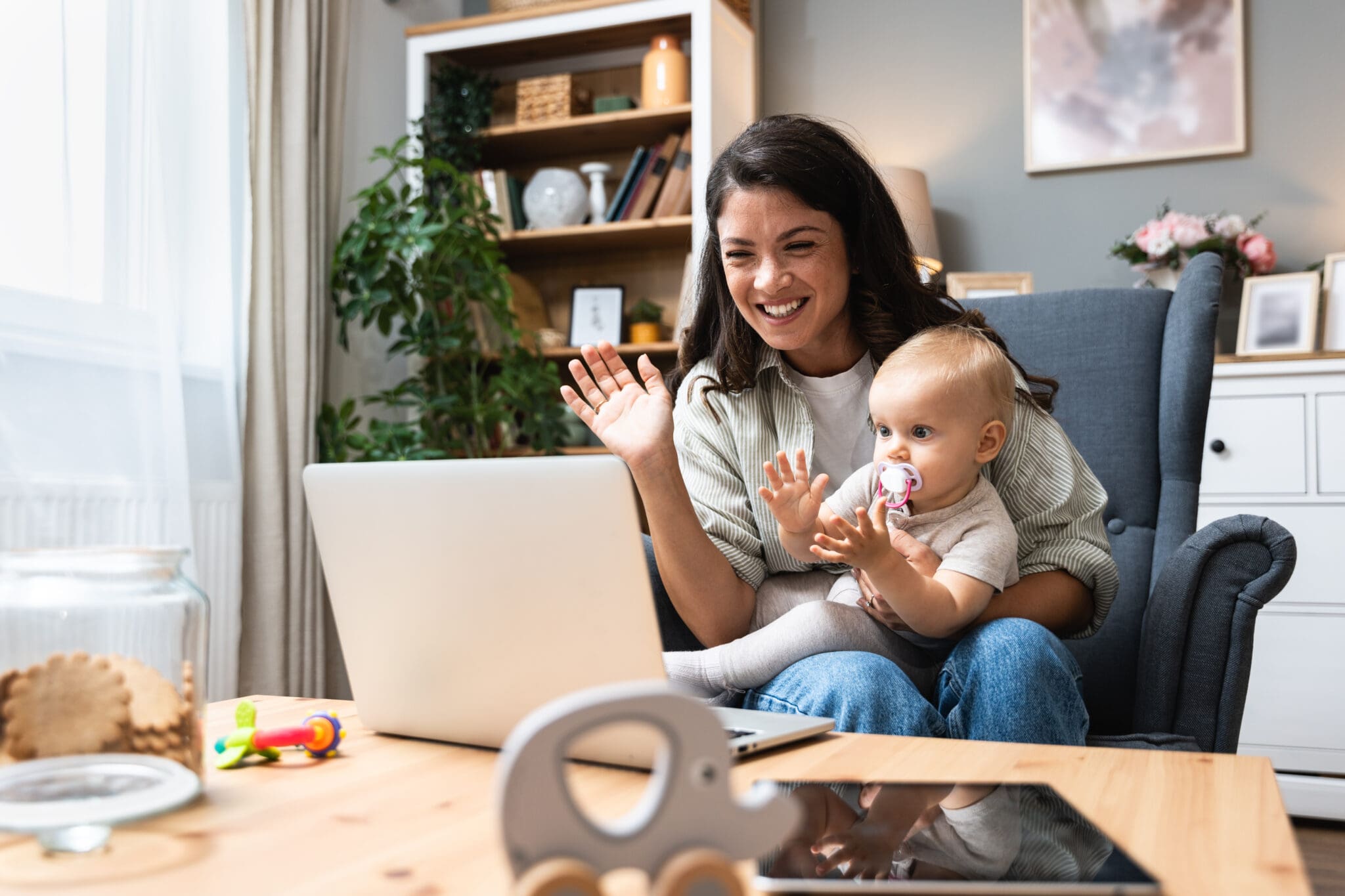Stay at home mom working remotely on laptop while taking care of her baby. Young business mother on maternity leave trying to freelance by the desk with toddler child. Global work on computer Woman and baby video calling on laptop.