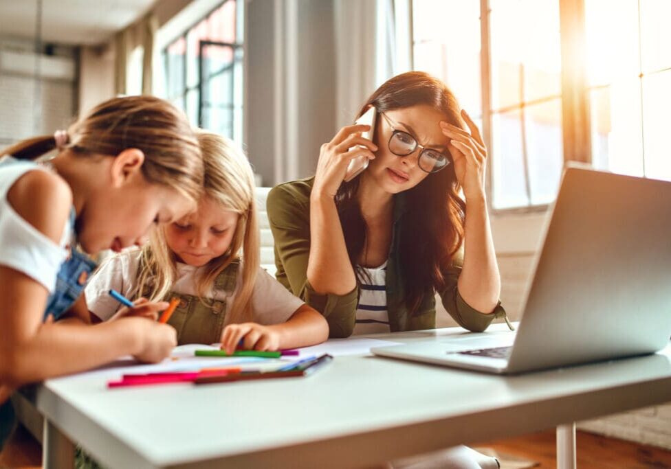 A business woman and a mother are trying to work on a laptop when her little daughters are playing, fooling around and interfering with her. Freelance, work from home. Mother stressed dealing with life and children while spouse is a away working.