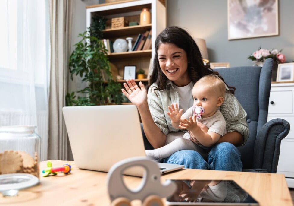 Stay at home mom working remotely on laptop while taking care of her baby. Young business mother on maternity leave trying to freelance by the desk with toddler child. Global work on computer Woman and baby video calling on laptop.