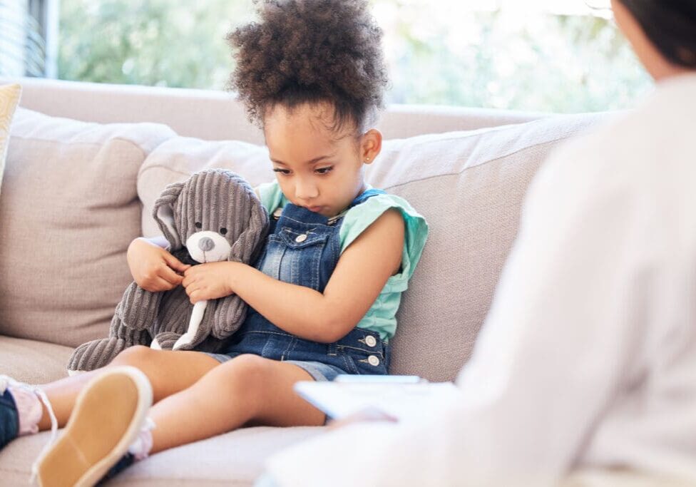 Child, sad and therapy in clinic for mental health, depression and conversation in doctors office. Young, girl and teddy bear for support, psychology and wellness for trauma on couch with therapist Child holding teddy bear on couch.