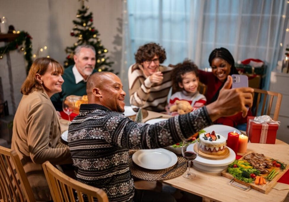 Multi-ethnic family using mobile phone video call online to relative. Attractive diverse group of people having dinner eating food to celebrate holiday Thanksgiving, X-mas eve on dining table at home.